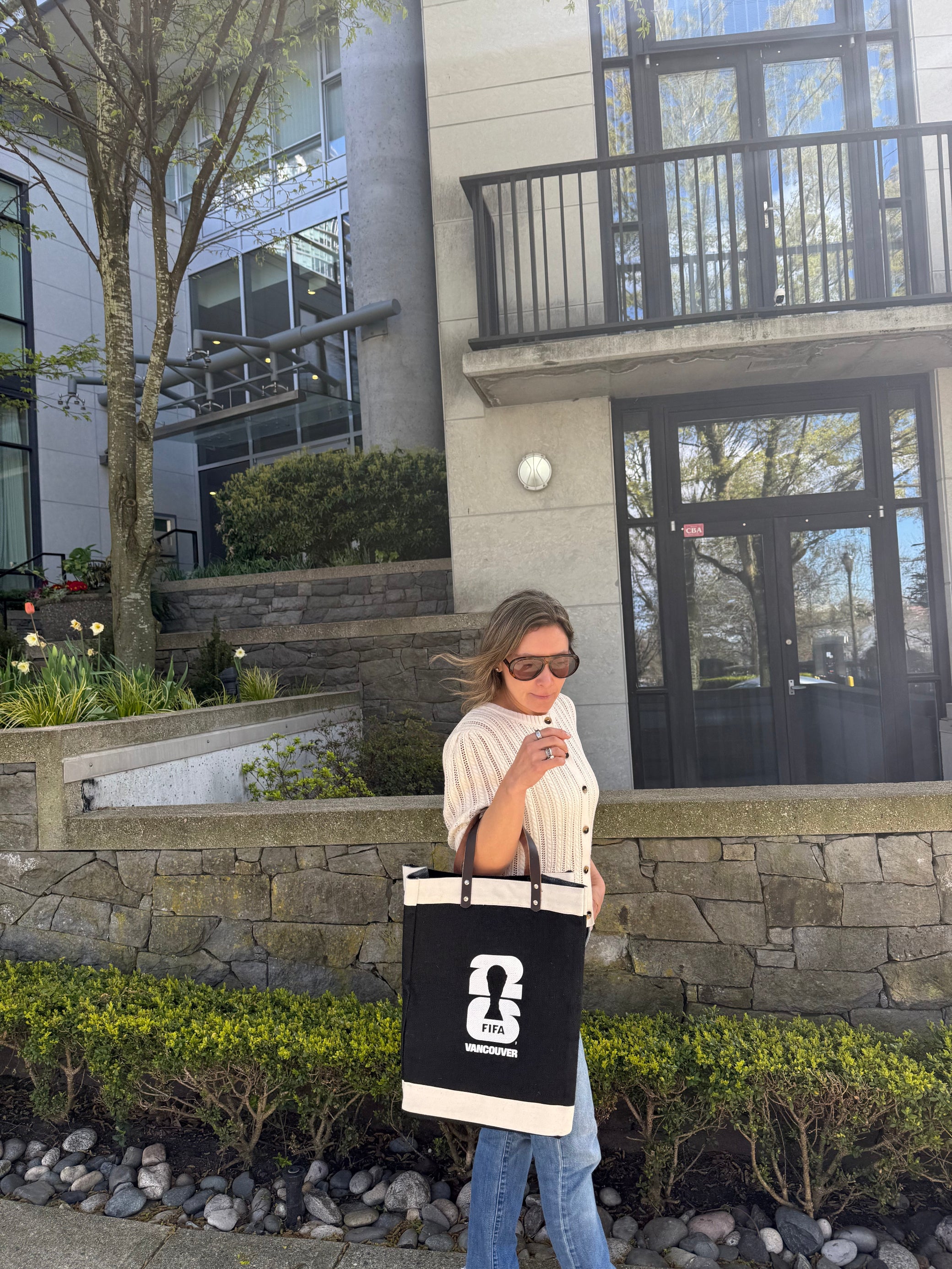 Person holding a large market bag with a FIFA Vancouver logo in front of a modern building.