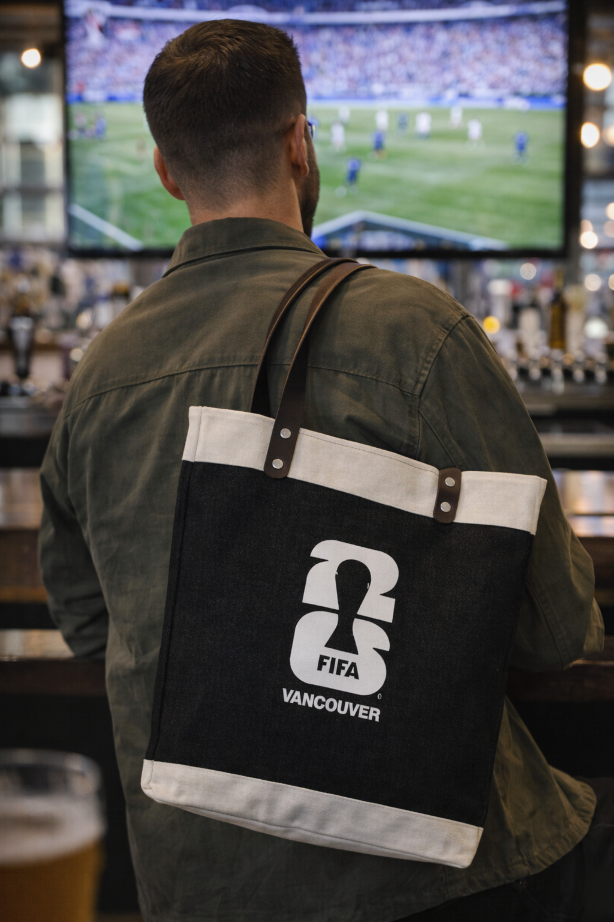 Person wearing a FIFA Vancouver tote market bag in a bar setting with sports on TV.