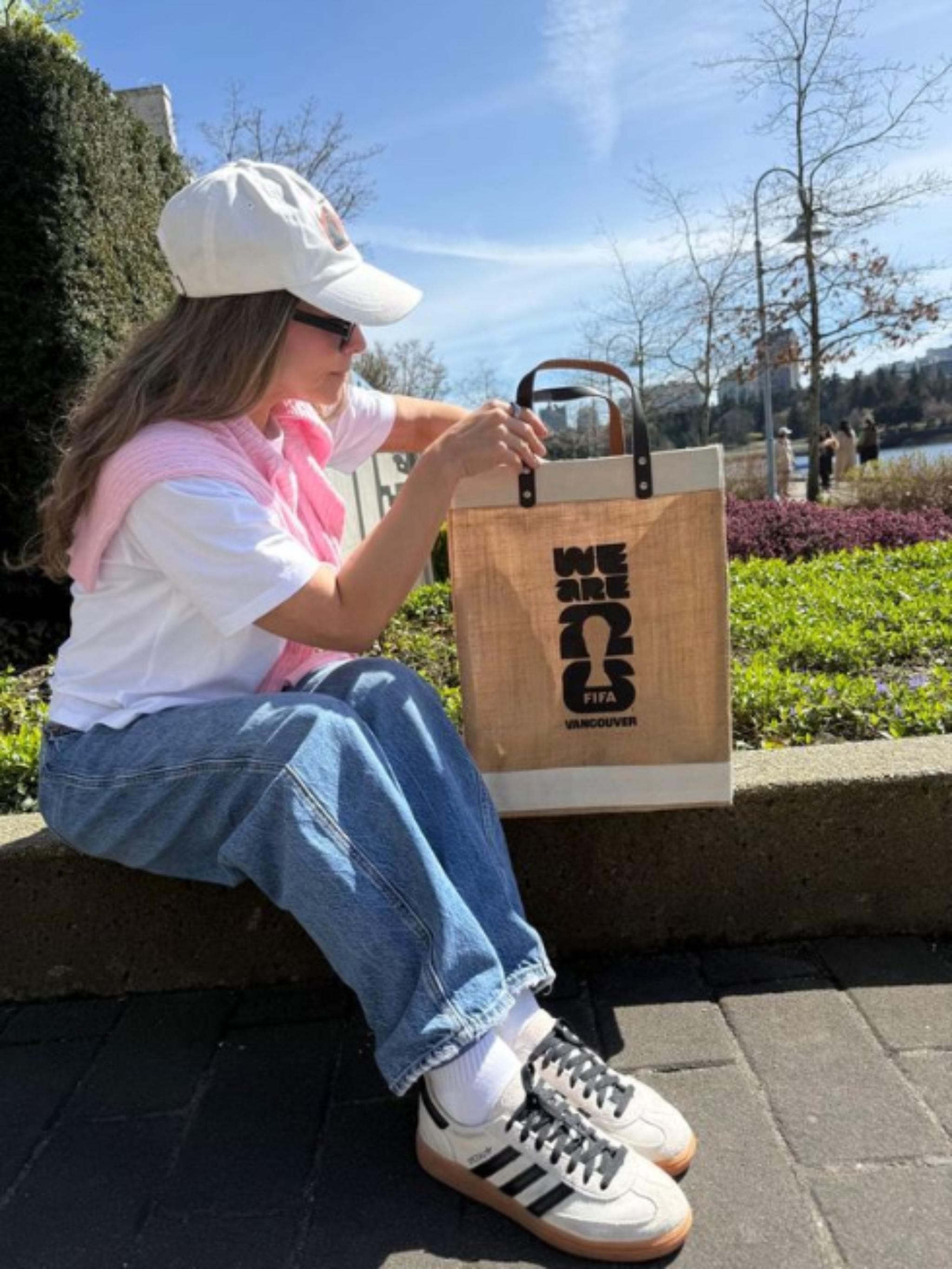 Person sitting outdoors with a market tote bag, wearing a white cap and jeans.